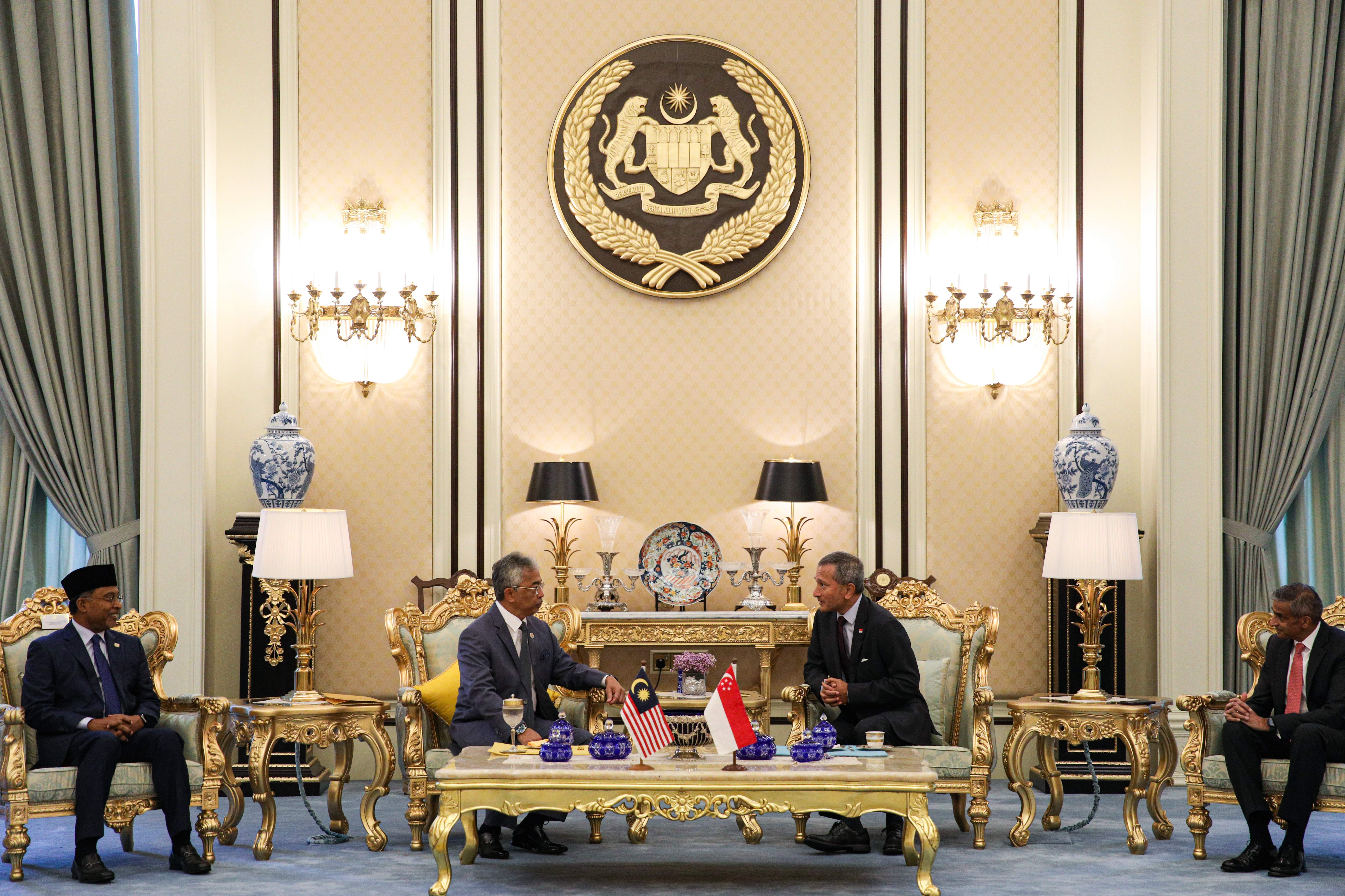Four men in suits sit in ornate chairs in a formal room with Malaysia's coat of arms.